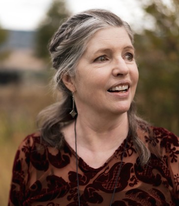 A middle-aged woman with gray hair, smiling while looking off to the side, standing outdoors with a blurred natural background.