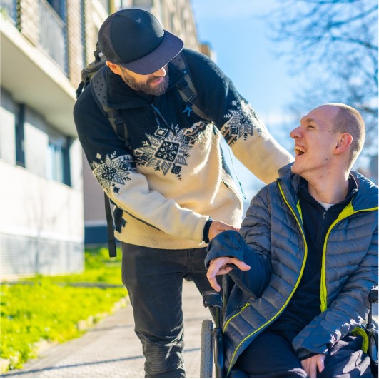 A man in a winter sweater is smiling and interacting warmly with a person in a wheelchair on a sunny day outside a building.